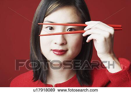 Stock Photo - Portrait of a young woman holding chopsticks in front of her face. Fotosearch