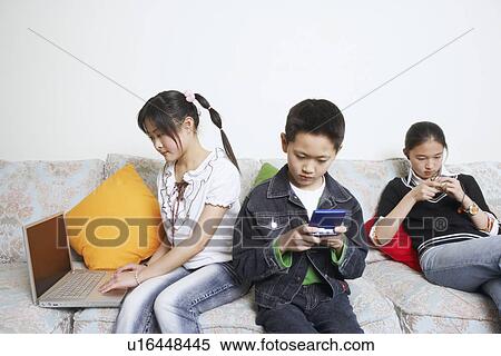 Boy sitting on a couch playing a video game beside his two sisters View Large Photo Image Stock Photography - Boy sitting on a couch playing a video game beside his two sisters. Fotosearch