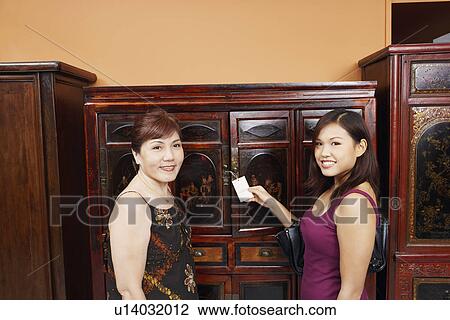 Stock Image - Portrait of a mature woman and her daughter smiling in front of an almirah. Fotosearch