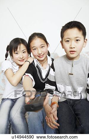 Stock Image - Portrait of a young woman sitting with her sister and her brother holding a remote control. Fotosearch