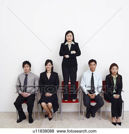 Stock Photo - Portrait of a businesswoman standing on an armchair with four business executives sitting beside her. Fotosearch