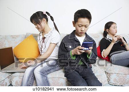 Stock Image - Boy sitting on a couch playing a video game beside his two sisters. Fotosearch