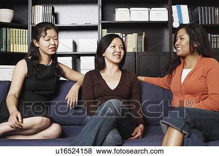 Stock Photo - Close-up of a mid adult woman sitting with two young women on a couch smiling. Fotosearch