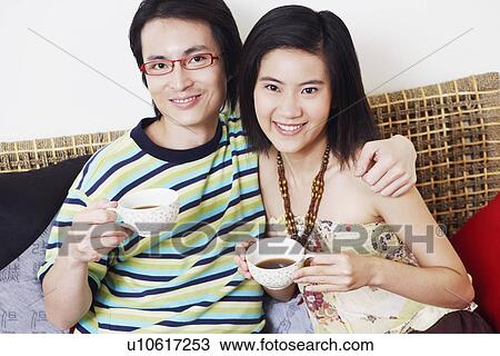 Stock Image - Portrait of a mid adult man and a young woman holding cups of tea and smiling. Fotosearch
