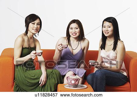 Stock Photo - Portrait of three young women sitting on a couch with tea cups. Fotosearch