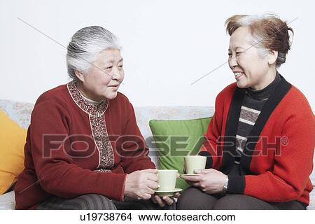 Picture - Side profile of a senior woman sitting on a couch with a mature woman holding tea cups. Fotosearch