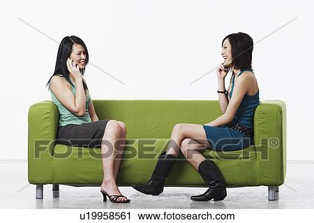 Stock Image - Side profile of two young women sitting on a couch talking on mobile phones. Fotosearch
