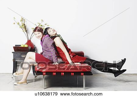 Stock Photo - Side profile of two young women sitting on a couch smiling. Fotosearch