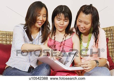 Stock Photo - Three young women sitting together on a couch and watching a photo album. Fotosearch