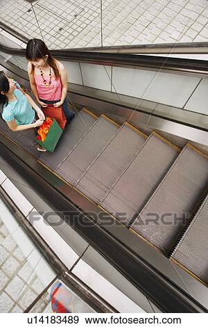 Stock Photo - High angle view of two young women standing on an escalator with shopping bags. Fotosearch