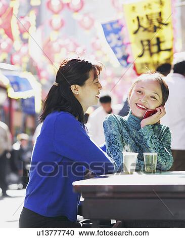 Stock Image - Young woman talking on a mobile phone sitting with another young woman in a restaurant. Fotosearch