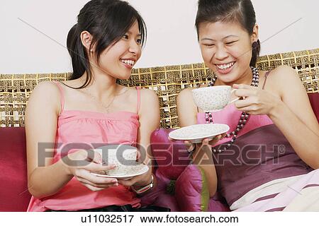 Stock Photo - Close-up of two young women sitting on a couch and holding tea cups. Fotosearch