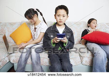 Picture - Portrait of a boy playing a video game beside his two sisters. Fotosearch