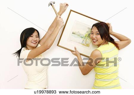 Stock Image - Portrait of two young women mounting paintings on a wall. Fotosearch