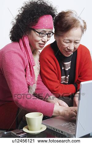 Stock Image - Side profile of a mature woman sitting with her friend using a laptop. Fotosearch