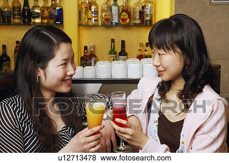 Stock Photo - Two young women sitting at a bar counter raising a toast. Fotosearch
