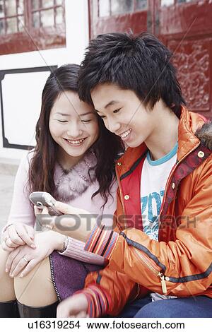 Picture - Teenage boy sitting together with a young woman holding a mobile phone. Fotosearch