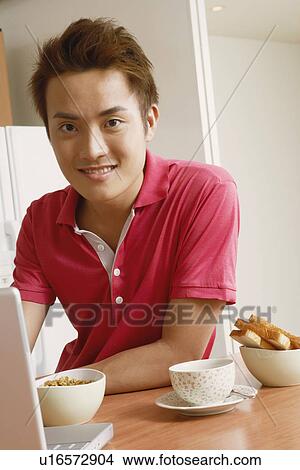 Picture - Portrait of a young man smiling in front of a laptop at a kitchen counter. Fotosearch