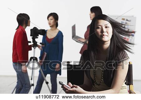 Portrait of a young woman holding a mobile phone with her friends standing behind her View Large Photo Image Stock Image - Portrait of a young woman holding a mobile phone with her friends standing behind her. Fotosearch