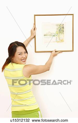 Stock Image - Portrait of a young woman hanging a picture frame on a wall and smiling. Fotosearch