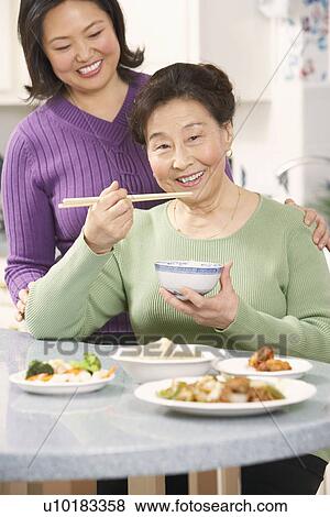 Portrait of a senior woman eating food with her daughter standing behind her View Large Photo Image Stock Photo - Portrait of a senior woman eating food with her daughter standing behind her. Fotosearch