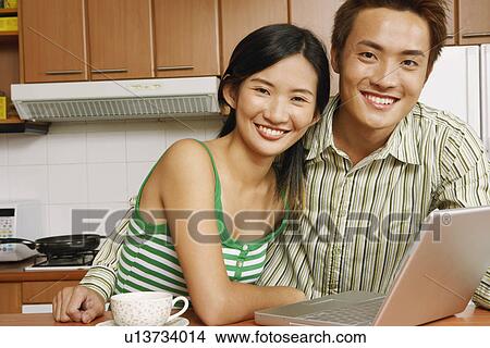 Picture - Portrait of a young couple smiling in front of a laptop at a kitchen counter. Fotosearch