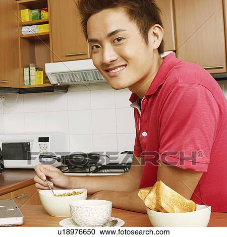 Stock Image - Portrait of a young man having breakfast in front of a laptop at a kitchen counter. Fotosearch