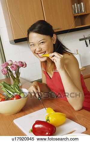 Stock Photography - Portrait of a young woman eating a slice of yellow bell pepper at a kitchen counter. Fotosearch