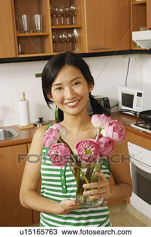 Stock Image - Portrait of a young woman holding a flower vase in the kitchen. Fotosearch