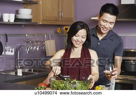 Young man standing holding a wineglass with a young woman preparing food View Large Photo Image Picture - Young man standing holding a wineglass with a young woman preparing food. Fotosearch