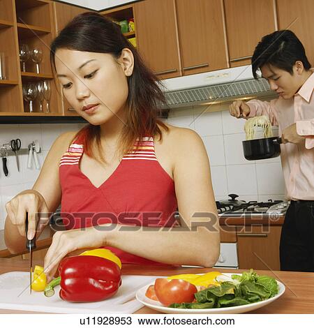 Young woman cutting vegetables in the kitchen with a young man eating noodles in the background View Large Photo Image Stock Image - Young woman cutting vegetables in the kitchen with a young man eating noodles in the background. Fotosearch