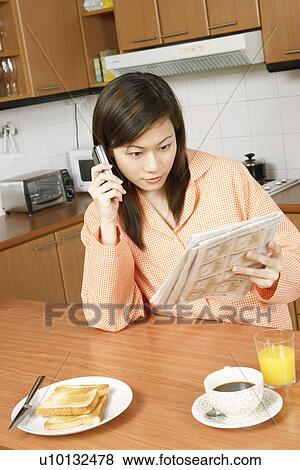 Young woman reading a newspaper and using a mobile phone at a kitchen counter View Large Photo Image Stock Photo - Young woman reading a newspaper and using a mobile phone at a kitchen counter. Fotosearch