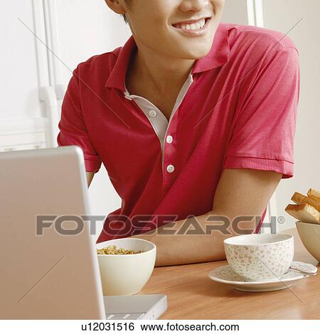 Stock Photograph - Close-up of a young man smiling in front of a laptop at a kitchen counter. Fotosearch