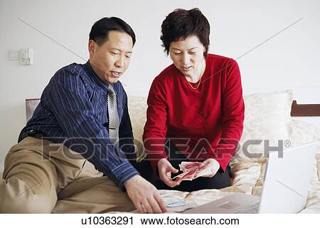 Stock Image - High angle view of a mature couple sitting on the bed with paper currency and a laptop. Fotosearch