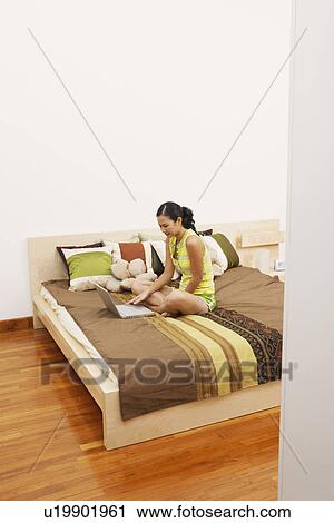 Stock Image - High angle view of a young woman sitting on the bed and using a laptop. Fotosearch