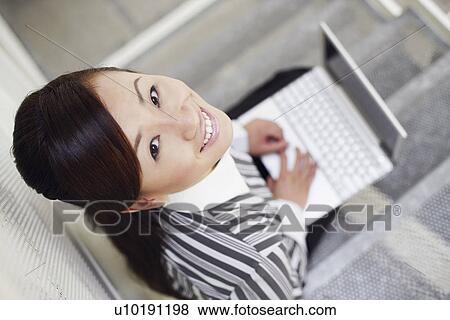 Stock Photo - High angle view of a young woman sitting on a staircase and using a laptop. Fotosearch