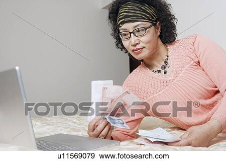 Stock Photo - Mature woman lying in front of a laptop holding paper currency. Fotosearch