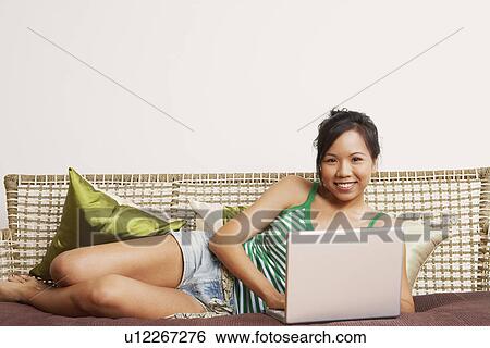 Portrait of a young woman lying on a couch and using a laptop View Large Photo Image Stock Photograph - Portrait of a young woman lying on a couch and using a laptop. Fotosearch