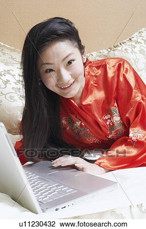 Stock Image - Portrait of a young woman lying on the bed with a laptop. Fotosearch