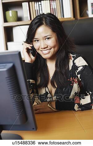 Stock Photograph - Portrait of a businesswoman sitting in front of a computer monitor using a mobile phone. Fotosearch
