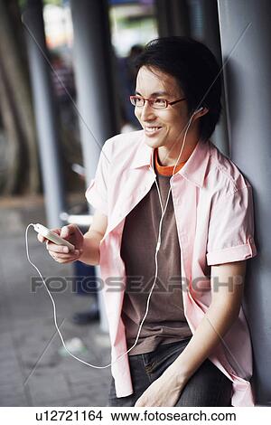 Picture - Close-up of a mid adult man leaning against a pole and listening to an MP3 player. Fotosearch