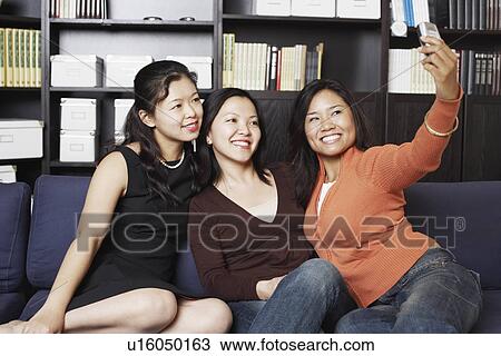 Close-up of a mid adult woman sitting with two young women on a couch taking a photograph of themselves View Large Photo Image Stock Image - Close-up of a mid adult woman sitting with two young women on a couch taking a photograph of themselves. Fotosearch