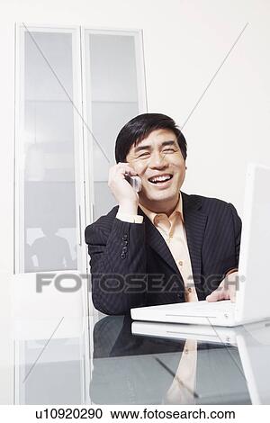 Stock Image - Businessman sitting in front of a laptop talking on a mobile phone. Fotosearch