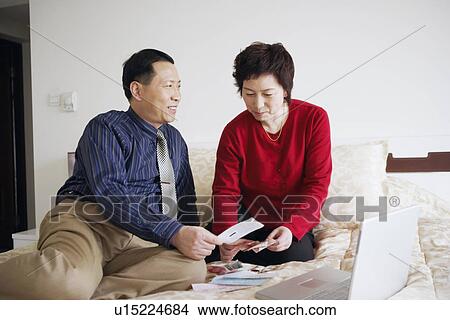 Picture - High angle view of a mature couple sitting on the bed with paper currency and a laptop. Fotosearch