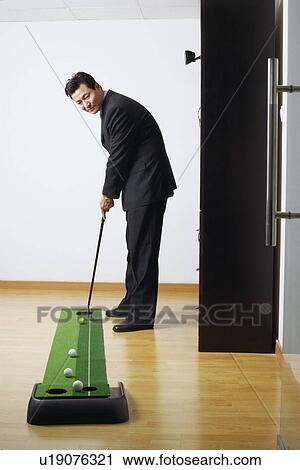 Stock Image - Side profile of a businessman practicing golf in a room. Fotosearch