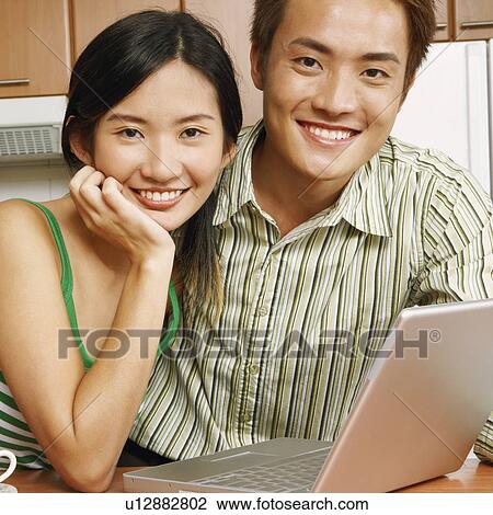 Stock Image - Portrait of a young couple smiling in front of a laptop at a kitchen counter. Fotosearch