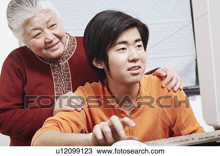 Stock Image - Close-up of a grandson using a computer with his grandmother standing behind him. Fotosearch