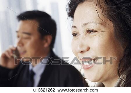 Close-up of a mature woman smiling with a mature man talking on a mobile phone behind her View Large Photo Image Stock Image - Close-up of a mature woman smiling with a mature man talking on a mobile phone behind her. Fotosearch