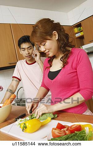 Picture - Close-up of a young woman cutting vegetables with a mid adult man talking on a mobile phone beside her. Fotosearch