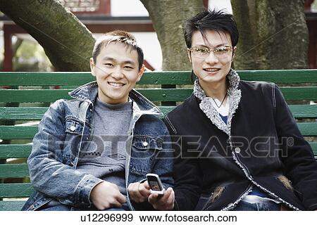 Portrait of a young man sitting together with his friend holding a mobile phone View Large Photo Image Stock Photo - Portrait of a young man sitting together with his friend holding a mobile phone. Fotosearch